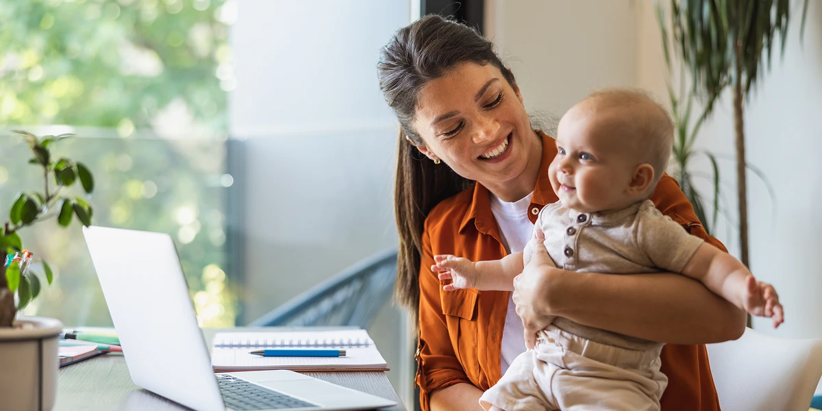Smiling mother holding her baby while working on a laptop at home