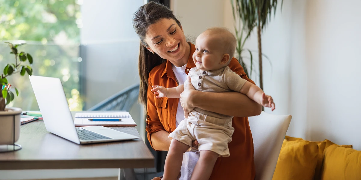 Smiling mother holding her baby while working on a laptop at home